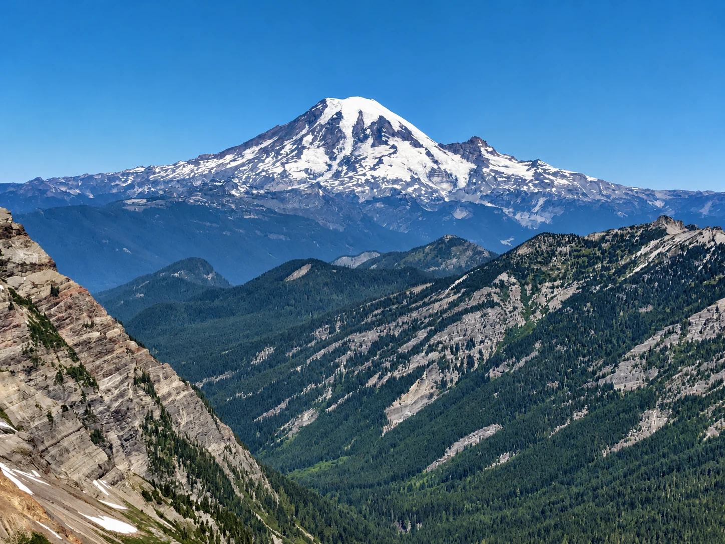 A snow-covered mountain above layered forested ridges under a clear blue sky
