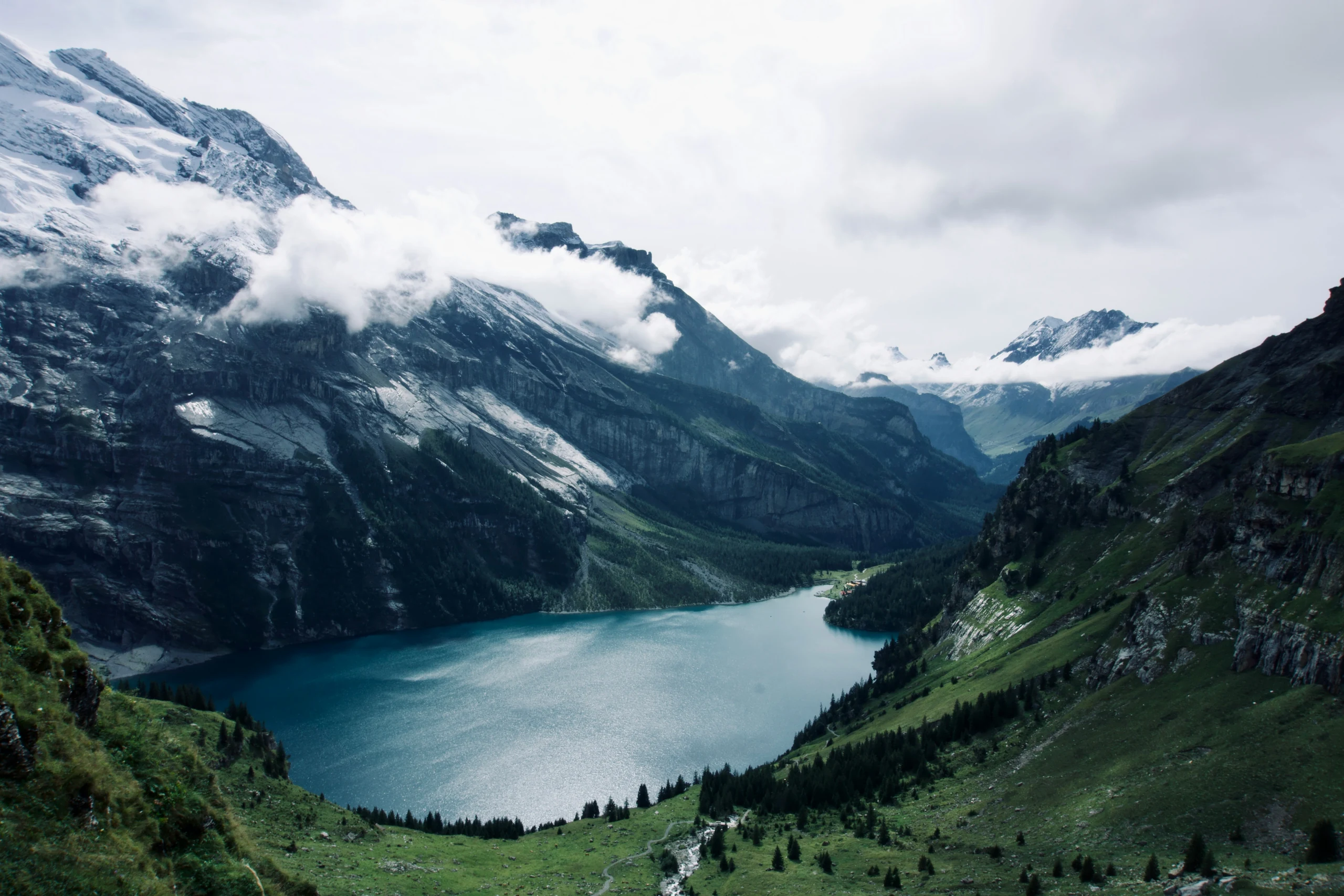 A blue alpine lake surrounded by forested slopes, snowy mountains, and low clouds